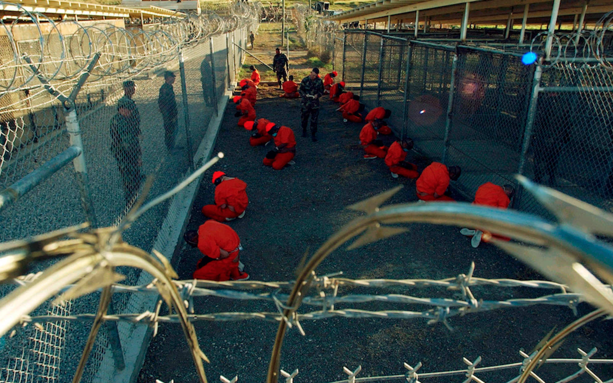 Detainees in orange jumpsuits sit in a holding area under the watchful eyes of Military Police at Camp X-Ray at Naval Base Guantanamo Bay, Cuba, during in-processing to the temporary detention facility on January 11, 2002. REUTERS/DoD/Shane T. McCoy