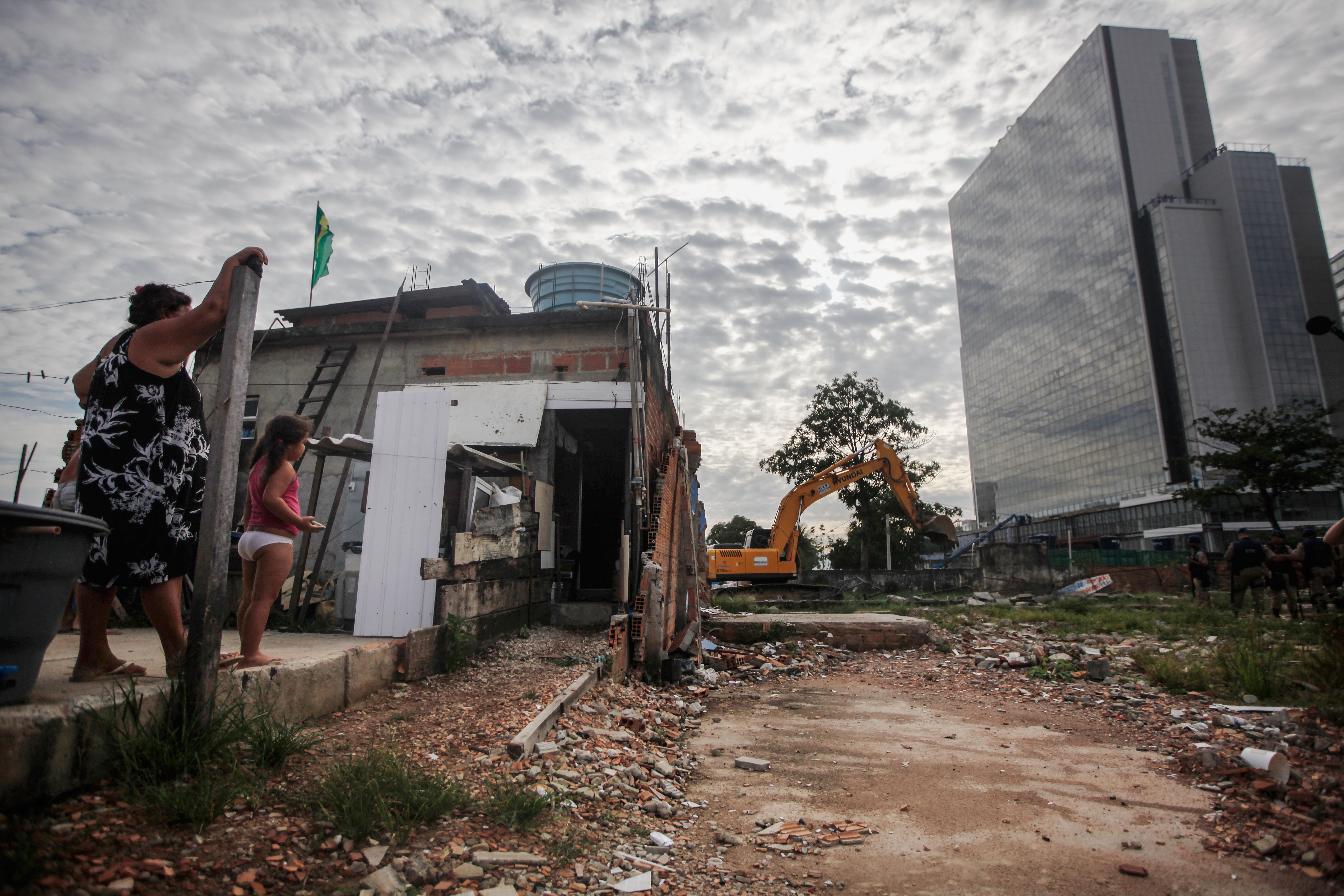 RIO DE JANEIRO, BRAZIL - FEBRUARY 24: Members of the Lopes family watch from their remaining home during the demolition of the neighboring residents association building in the mostly demolished Vila Autodromo favela community, with new Olympic Park construction standing (R), on February 24, 2016 in Rio de Janeiro, Brazil. Most residents of the favela community have moved out and had their properties demolished after receiving compensation for their homes which are located directly adjacent to the Olympic Park under construction for the Rio 2016 Olympic Games. A small fraction of remaining families from an original 700 or so in Vila Autodromo are resisting the controversial evictions and remain in the community. The favela sprang from an old fishing community and was considered one of the city's safest. Removals and demolitions have occurred in other Rio communities with tangential links to the games. (Photo by Mario Tama/Getty Images)